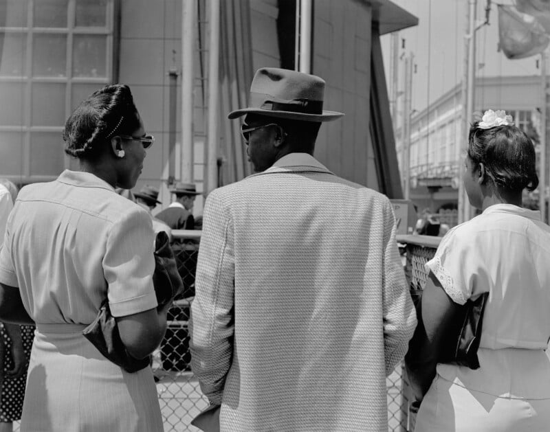 Three people stand with their backs to the camera, wearing mid-20th century attire. Two women and one man are engaged in conversation outdoors, near a fenced area and modern buildings.