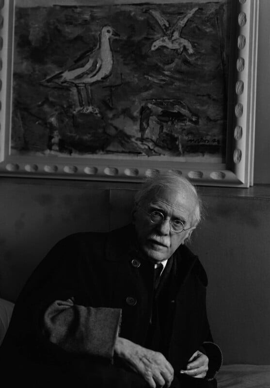 An elderly man with glasses and a mustache sits on a couch in front of a framed painting of birds. The image is in black and white, creating a contemplative and artistic atmosphere.