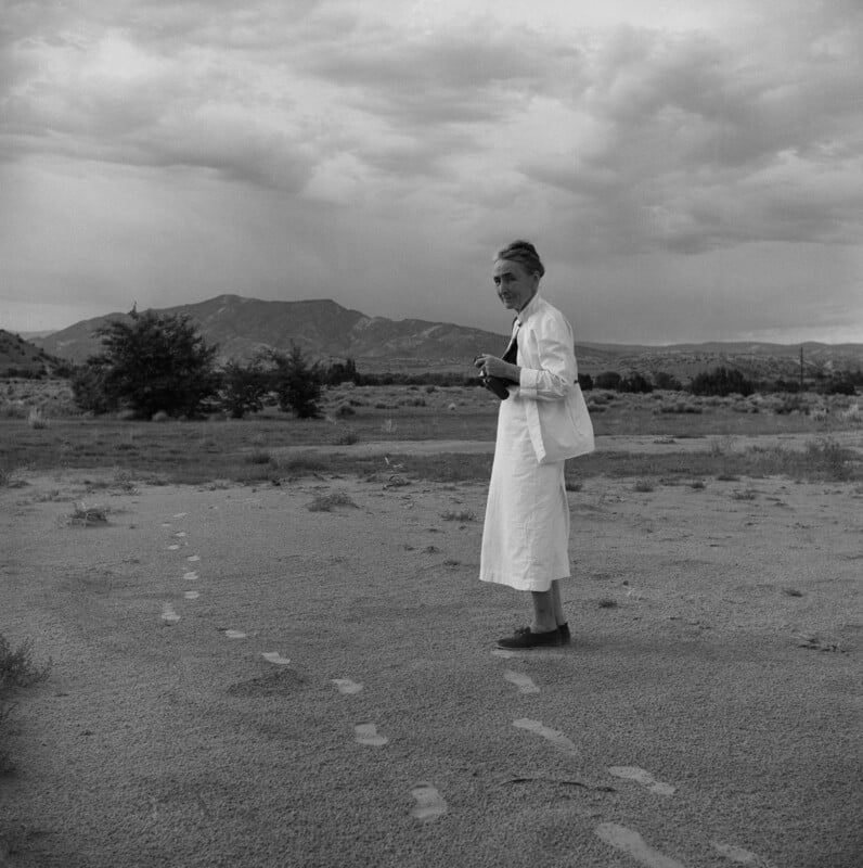 A woman in a long white dress stands on sandy ground with visible footprints, holding a camera, and looking back; mountains and cloudy skies appear in the background.