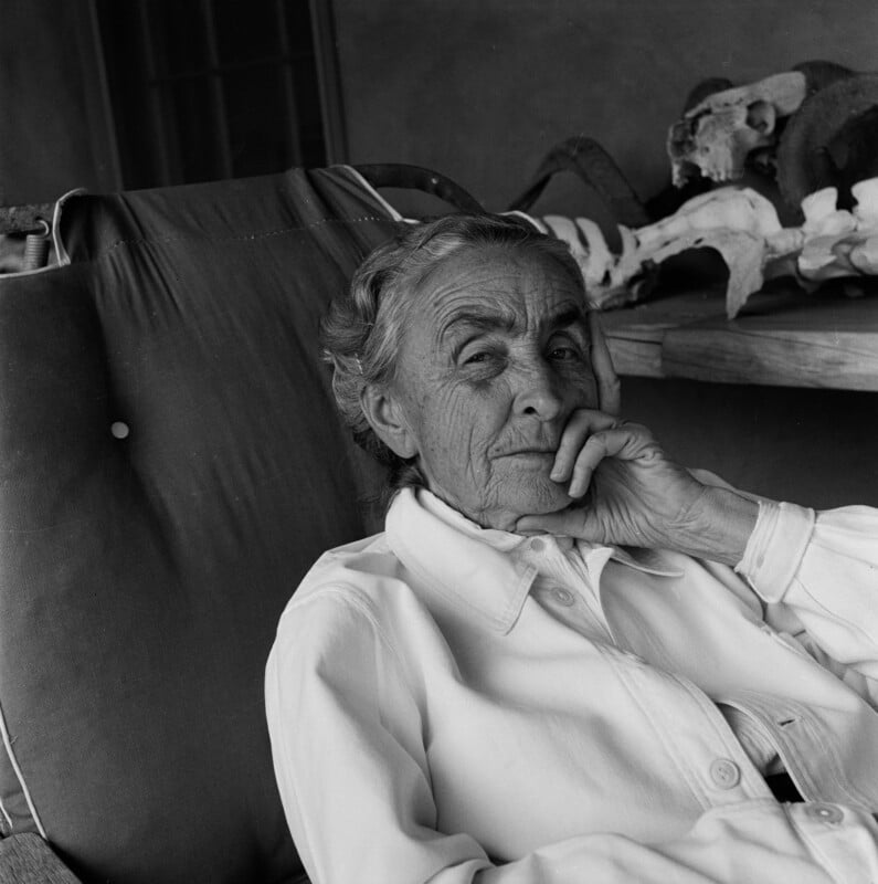 An older woman with gray hair sits in a cushioned chair, resting her chin on her hand and gazing thoughtfully. She wears a light-colored buttoned shirt. Animal bones are visible on a table behind her.