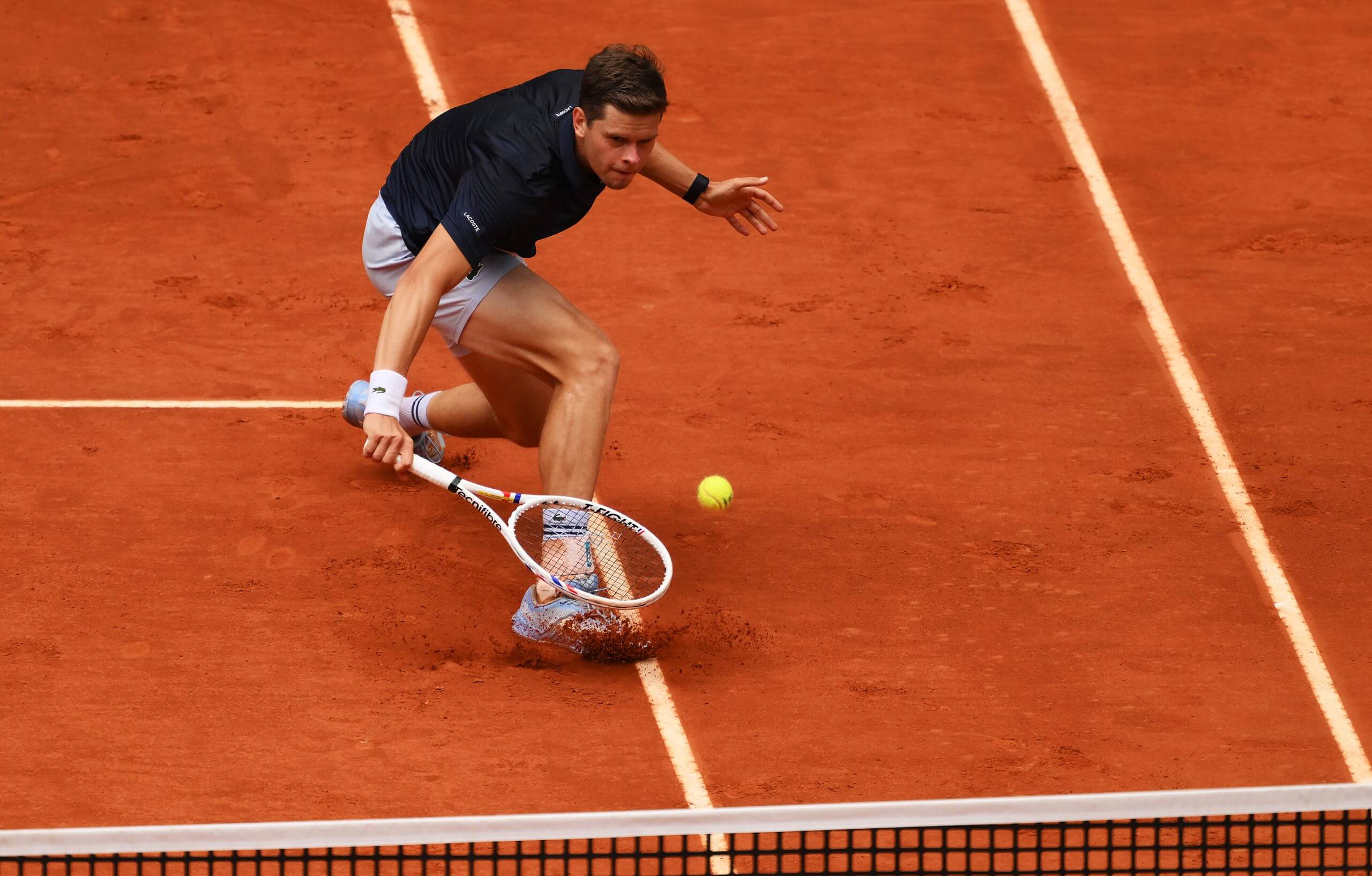 Alexander Blockx slides in to a backhand drop shot on a clay tennis court.