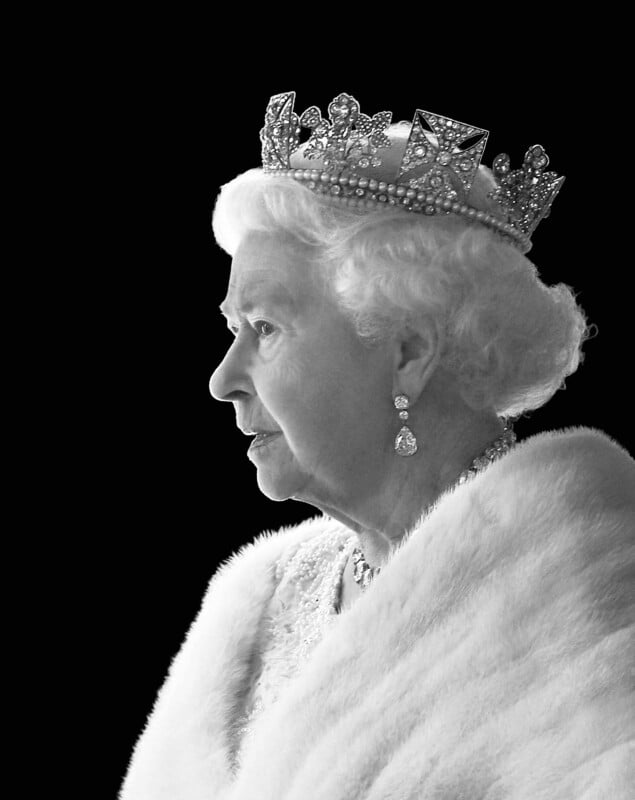 An elderly woman in profile wears a jeweled crown, earrings, and a fur-trimmed white garment against a black background. The image is black and white, highlighting her regal appearance.