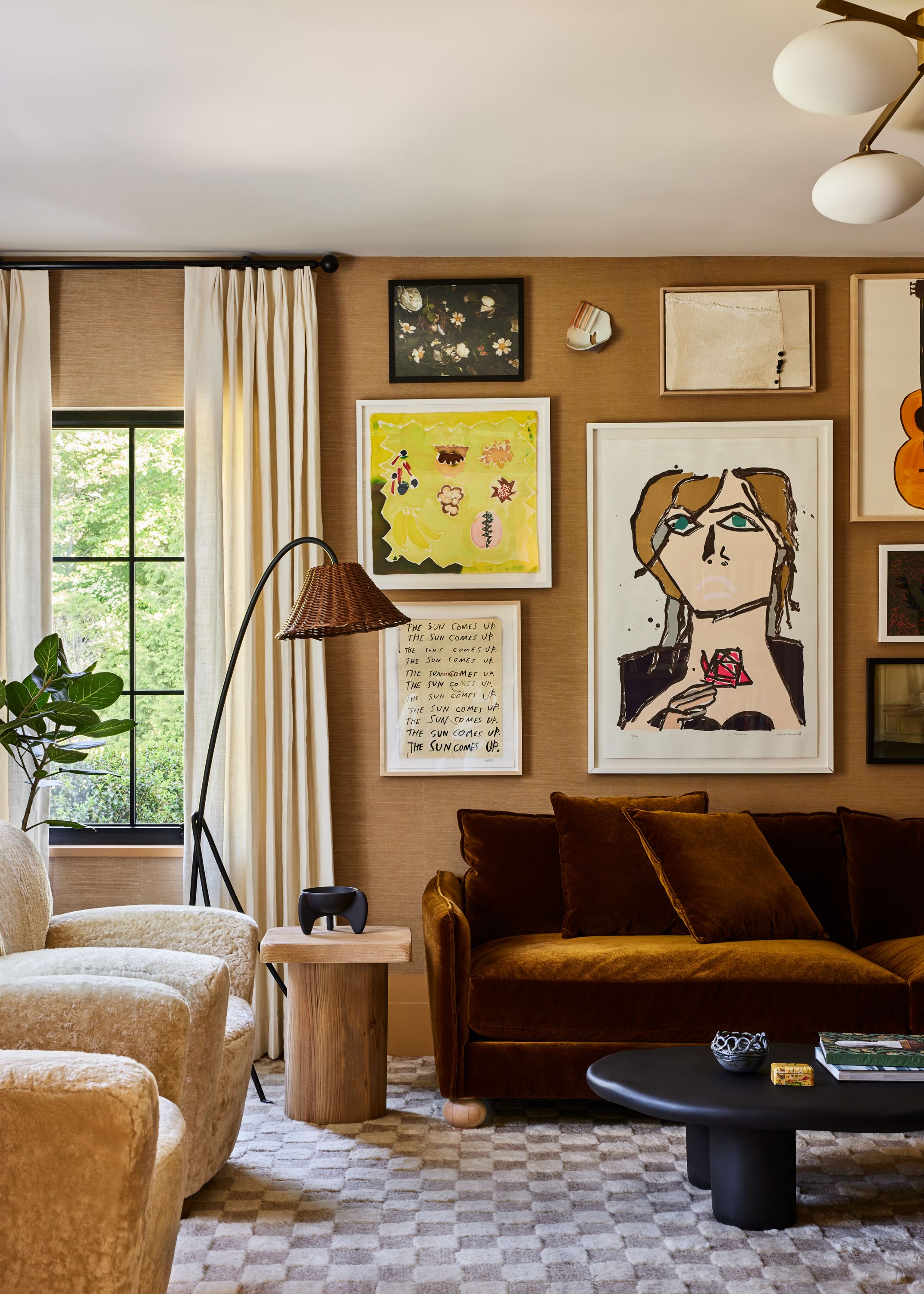 An earthy-toned, bright living room with a checkered, white and gray rug, upholstered armchairs and a sofa in beige fuzzy textile and brown velvet, and plenty of framed artworks hanging on the walls.