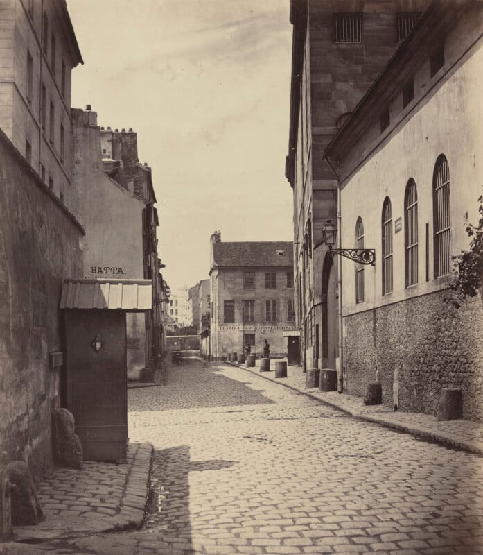 A black-and-white photo of a quiet, empty cobblestone street in a historic European city, lined with old stone buildings and shuttered windows under a partly cloudy sky.