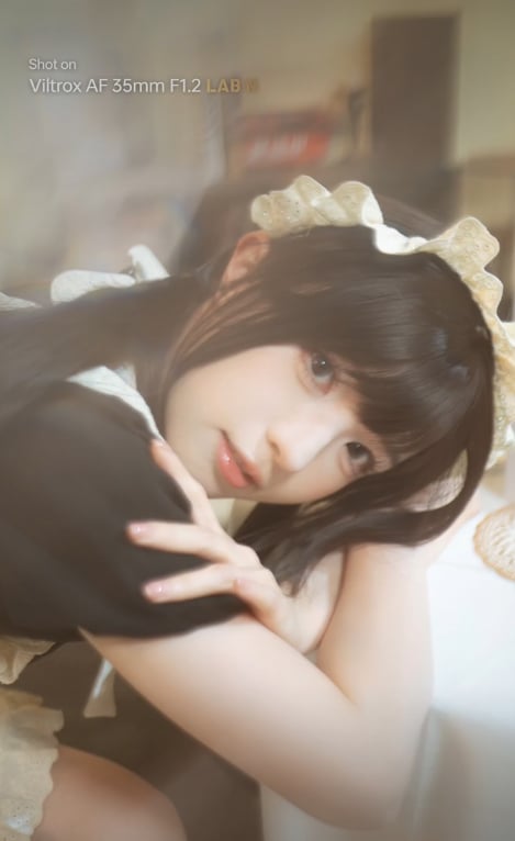 A person in a black and white maid outfit with a lace headband rests their arms on a table, gazing softly at the camera in a softly lit, blurred background.