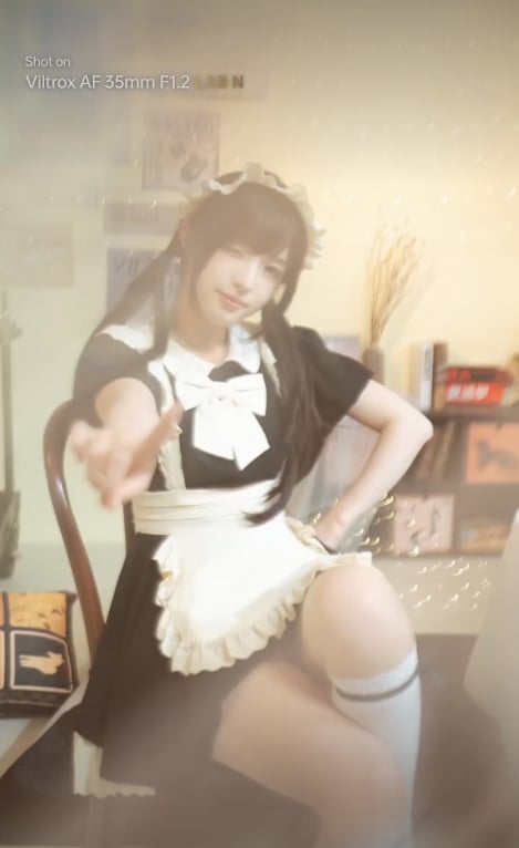 A person dressed in a black and white maid outfit poses while sitting on a chair, extending their hand toward the camera. The background features a cozy, softly lit room with books, pictures, and fairy lights.