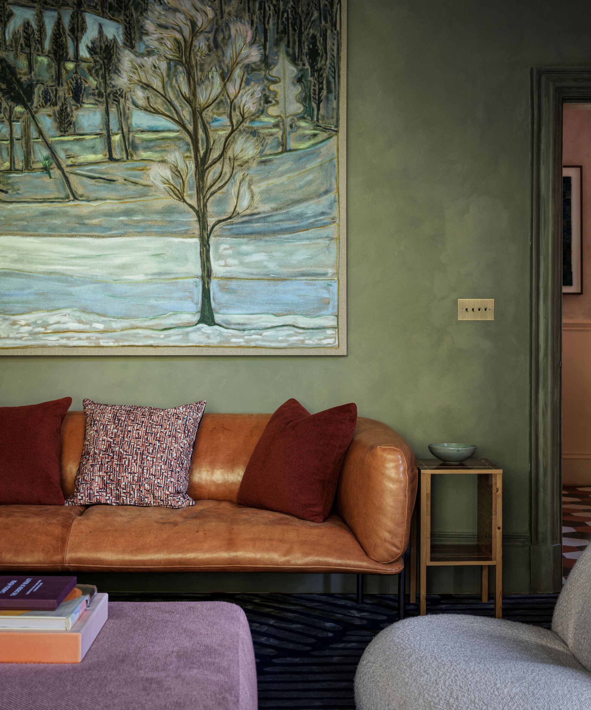 Living room with green limewash walls, a brown leather sofa with red cushions, a landscape painting of a forest, a pink ottoman, and a wooden side table with a blue bowl on top