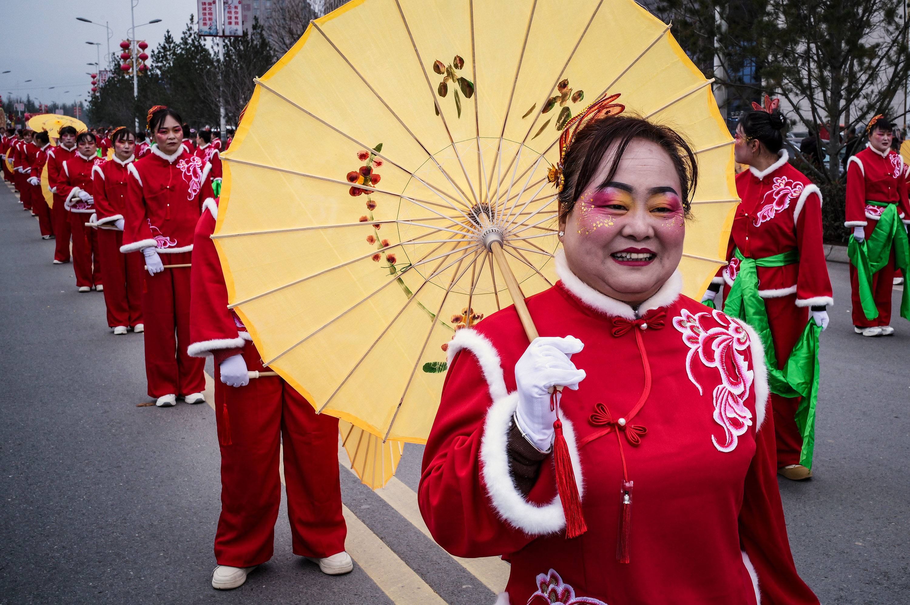 A woman in red costume holding a large yellow oil-paper umbrella leads a street procession with other costumed performers in matching red and green attire.