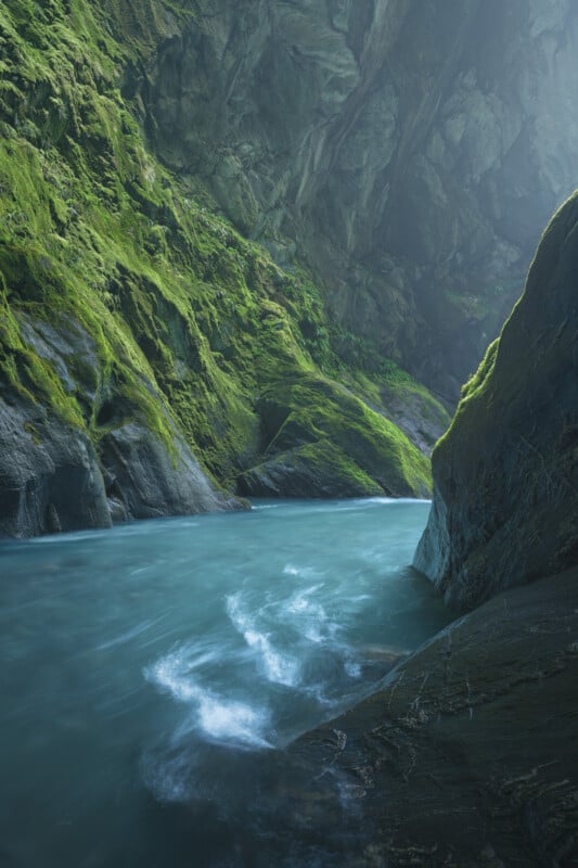 A tranquil blue river flows through a steep, narrow gorge with moss-covered rocky walls, bathed in soft, natural light from above.