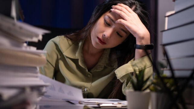 A woman in a green shirt looking tired, with a lot of work on her desk