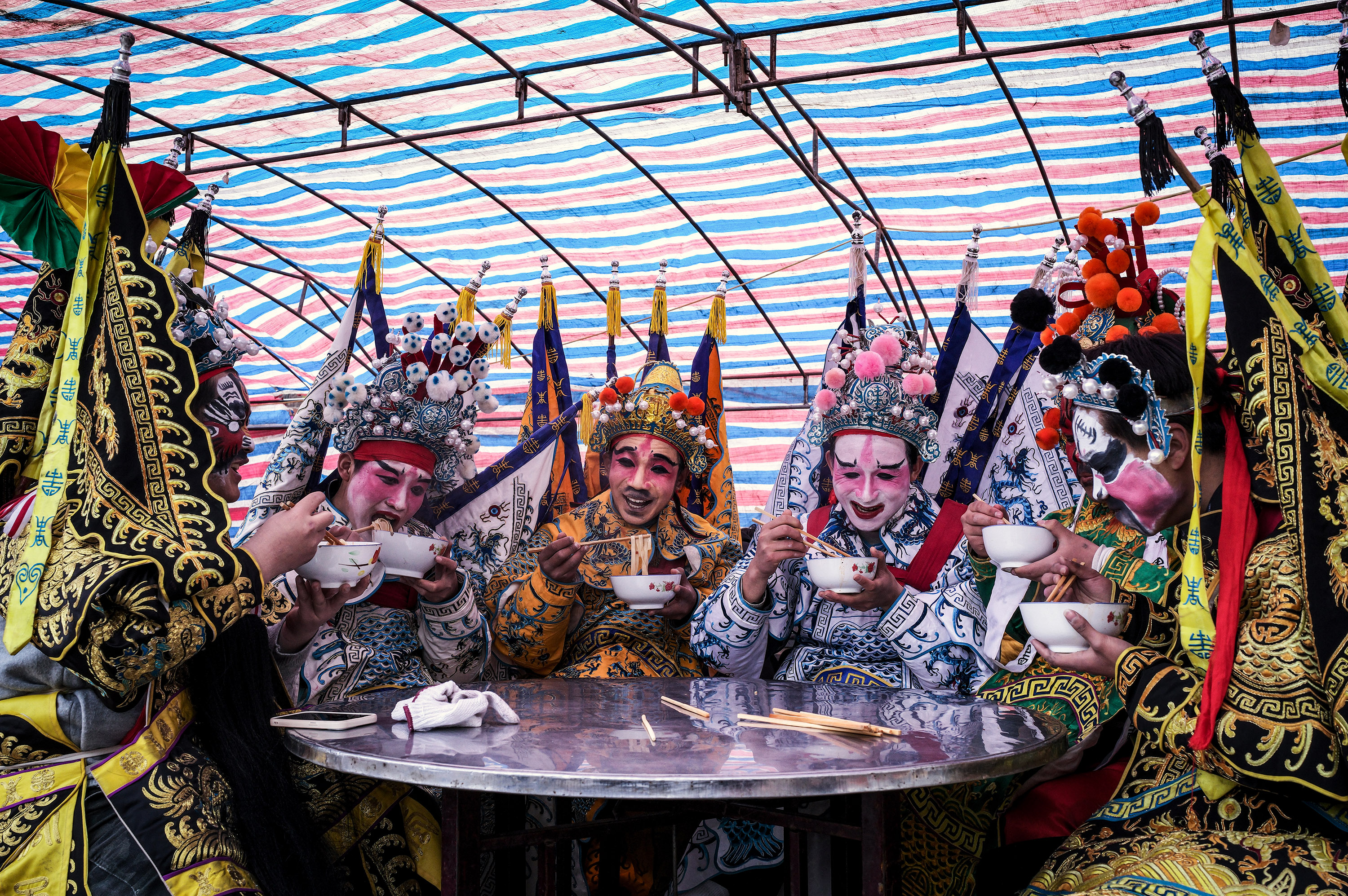 Performers in colorful traditional costumes and ornate headdresses eat from bowls at a round table beneath a striped tent and hanging red lanterns.