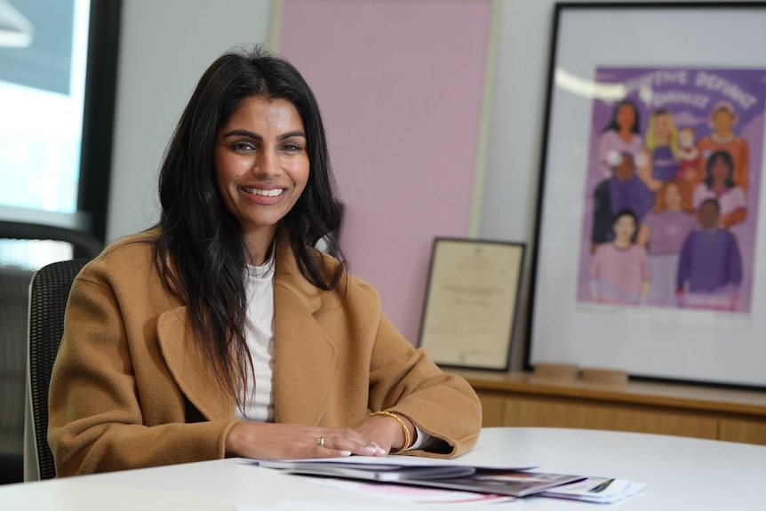 A young woman wearing a tan coat sitting at a table and smiling at the camera.