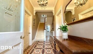A hallway into the crisis house - before renovations started - featuring Victorian-patterned brown floor tiles, a frosted glass door, stained wooden furniture, a white staircase and multiple chandeliers.