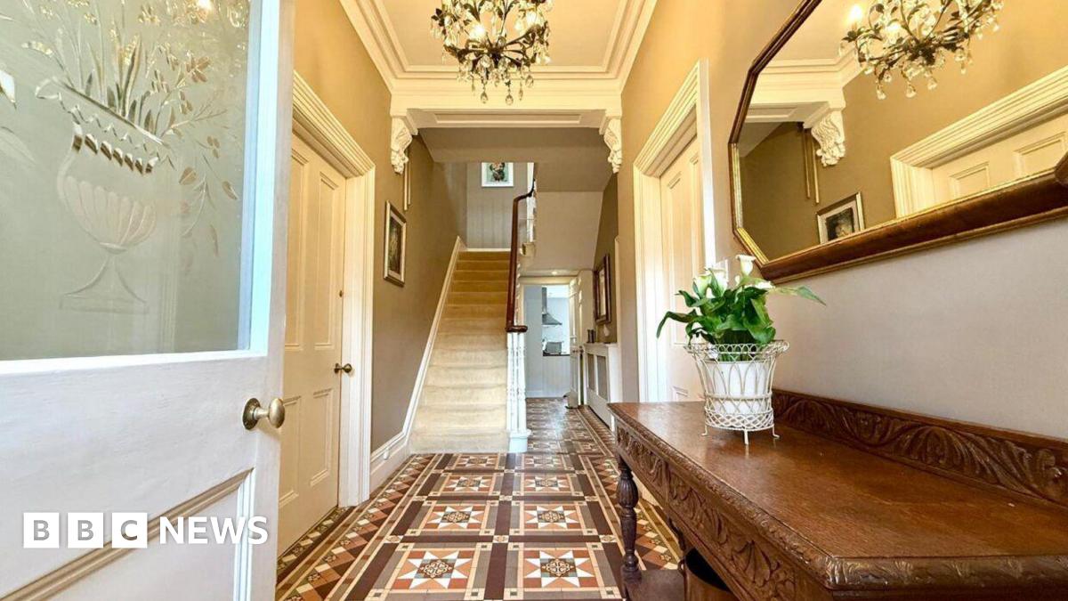 A hallway into the crisis house - before renovations started - featuring Victorian-patterned brown floor tiles, a frosted glass door, stained wooden furniture, a white staircase and multiple chandeliers.