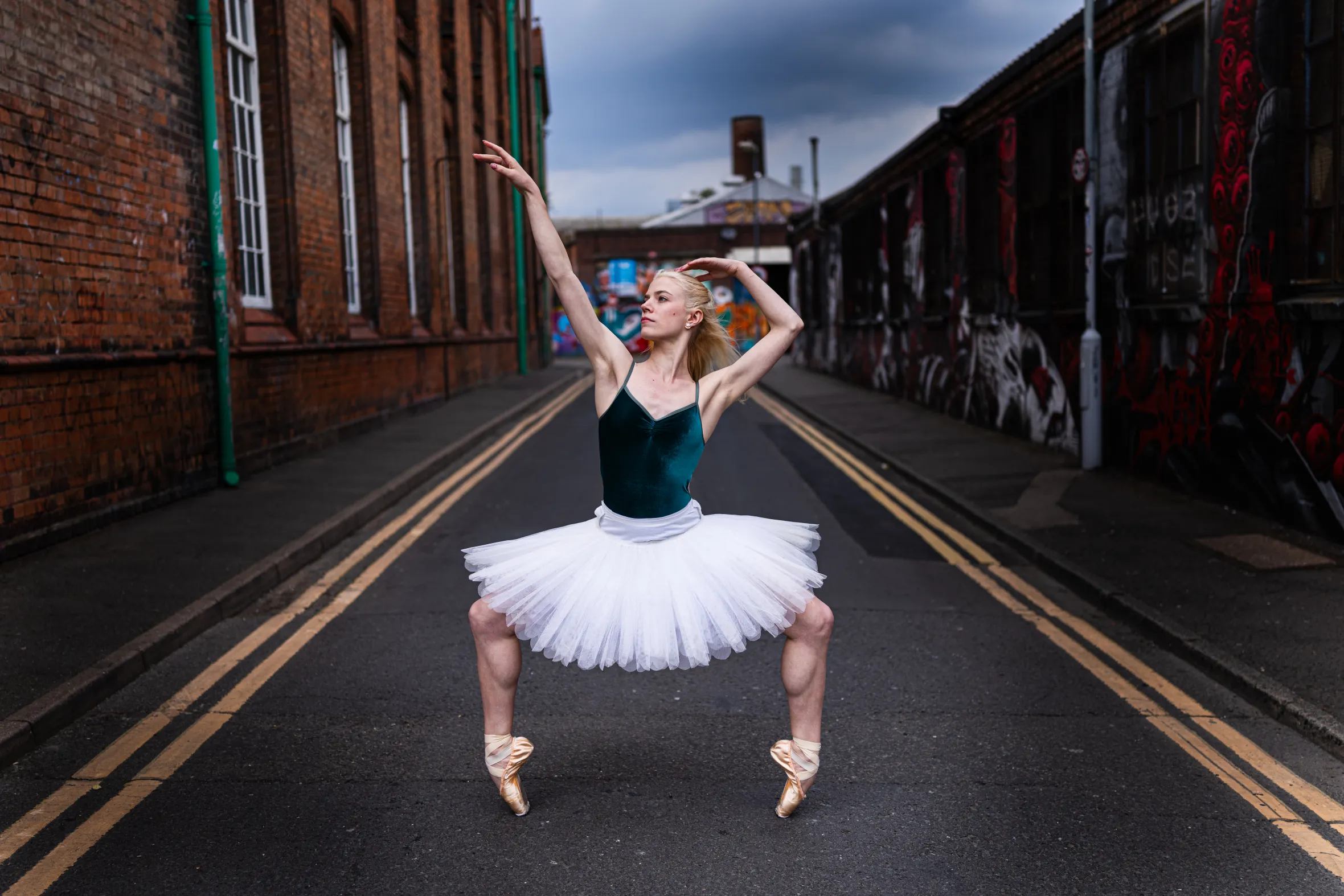 Birmingham Royal Ballet dancer Lucy Waine in a dark green leotard and white tutu, posing en pointe in the middle of a street between brick buildings.