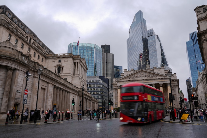 Sandstone buildings line a street with a red bus driving down the street and sky-scrapper buildings in the background