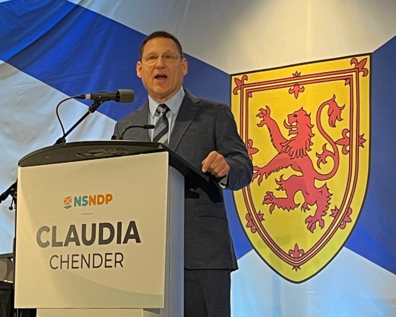 A middle aged bespectacled white man wearing a dark grey suit and pale shirt with blue and grey striped tie stands in front of a Nova Scotia flag. On the rostrum in front of him is a sign that says "NSNDP Claudia Chender."