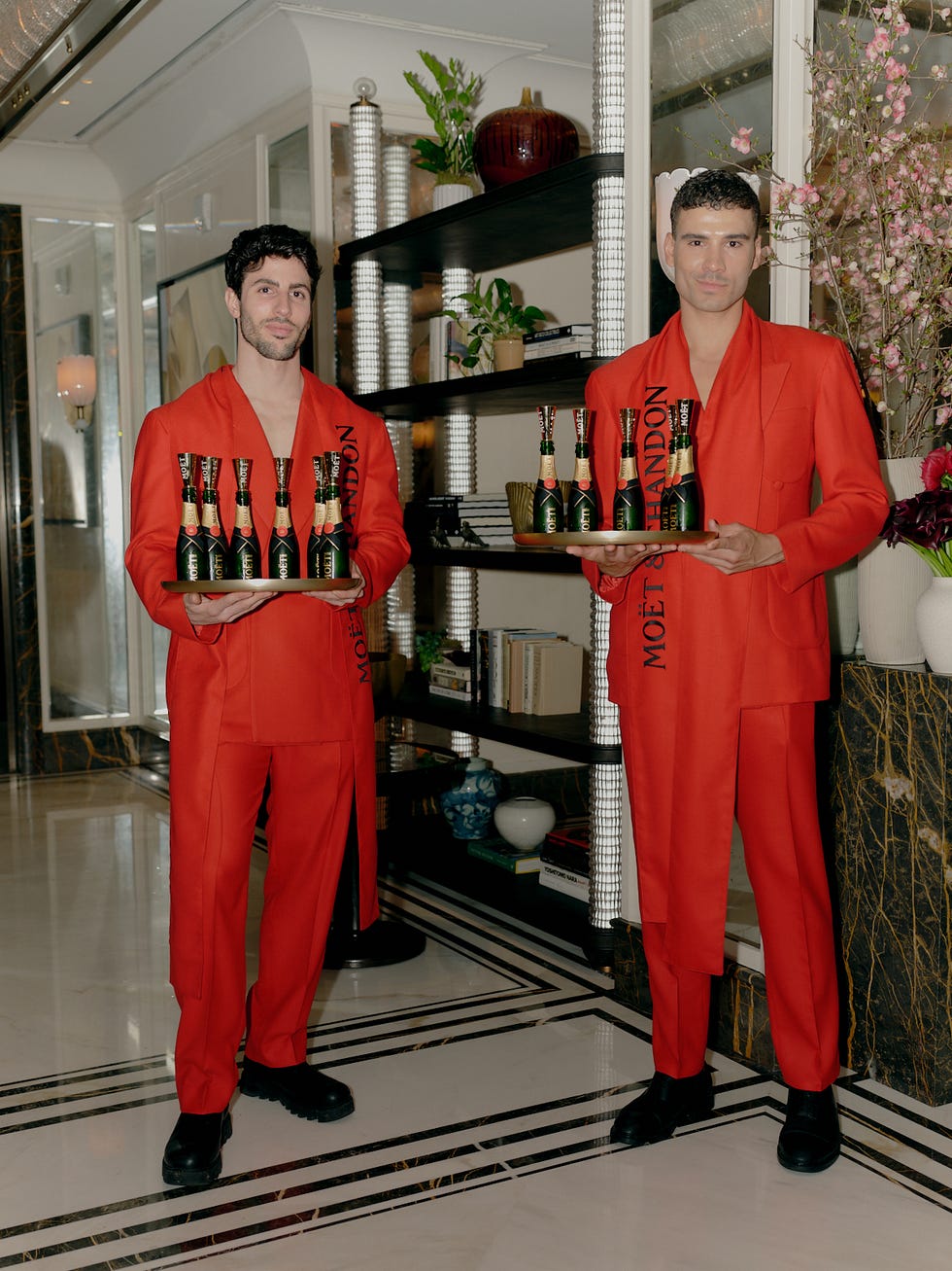 Two individuals holding trays of champagne bottles, dressed in matching red outfits.
