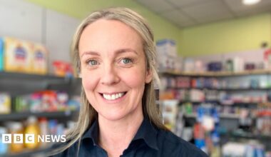 Two Female Pharmacists Working at a Pharmacy. They are wearing white coats and there are shelves filled with medication in the background.
