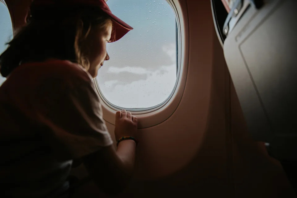 A little girl wearing a baseball cap embarks on a holiday. She sits at a window seat on a commercial airplane, and looks out at the clouds.