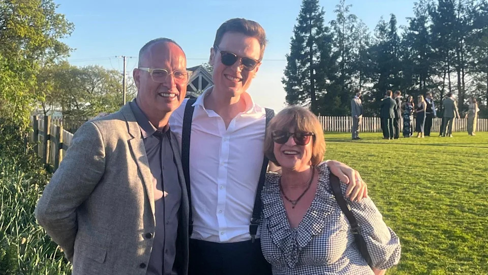 Three people stand smiling in a sunny field. Arthur's father is seen in a grey blazer on the left, Arthur is pictured in a white shirt and braces in the centre, and his mother is seen in a patterned dress on the right.