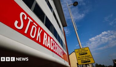 Outside of the STōK Racecourse. A red banner is across the building which reads 'STōK Racecourse' in capital letters. The font is bold and white. A lamp post can be seen next to the building which has a yellow and black coach drop-off sign on it.