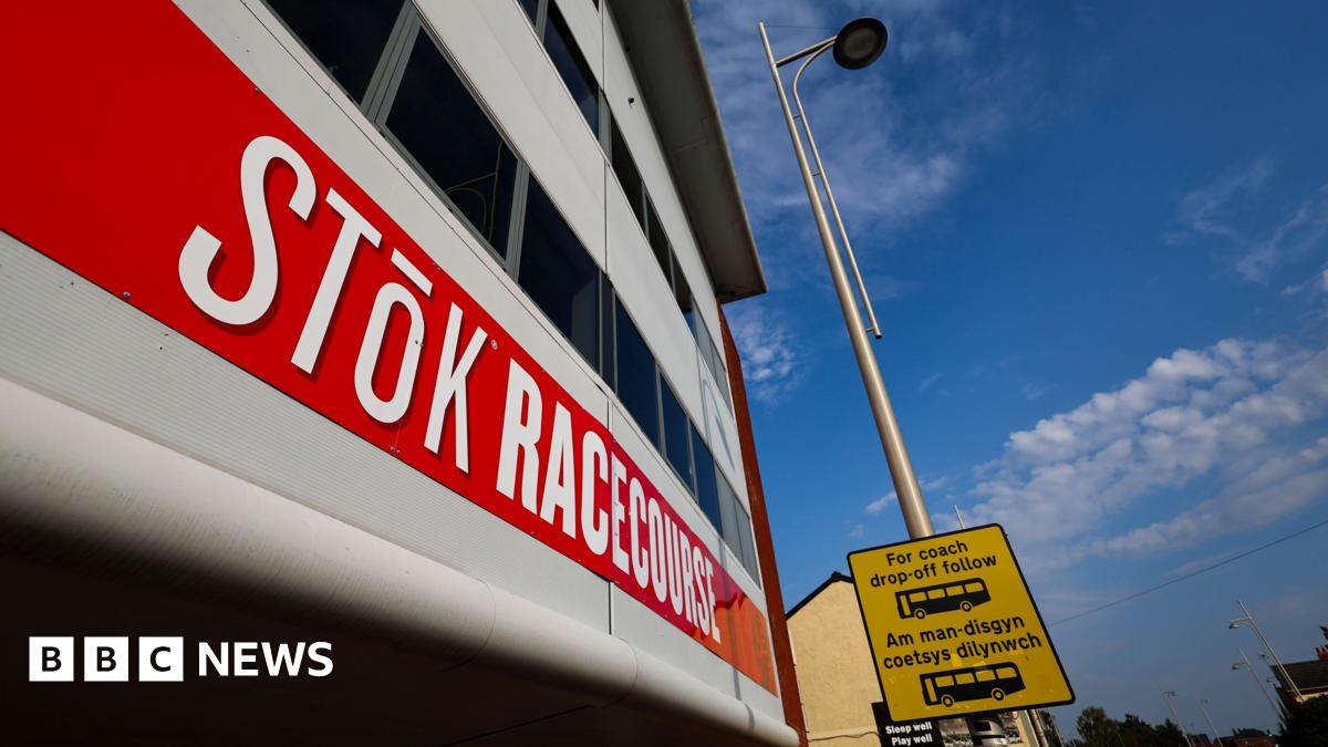 Outside of the STōK Racecourse. A red banner is across the building which reads 'STōK Racecourse' in capital letters. The font is bold and white. A lamp post can be seen next to the building which has a yellow and black coach drop-off sign on it.