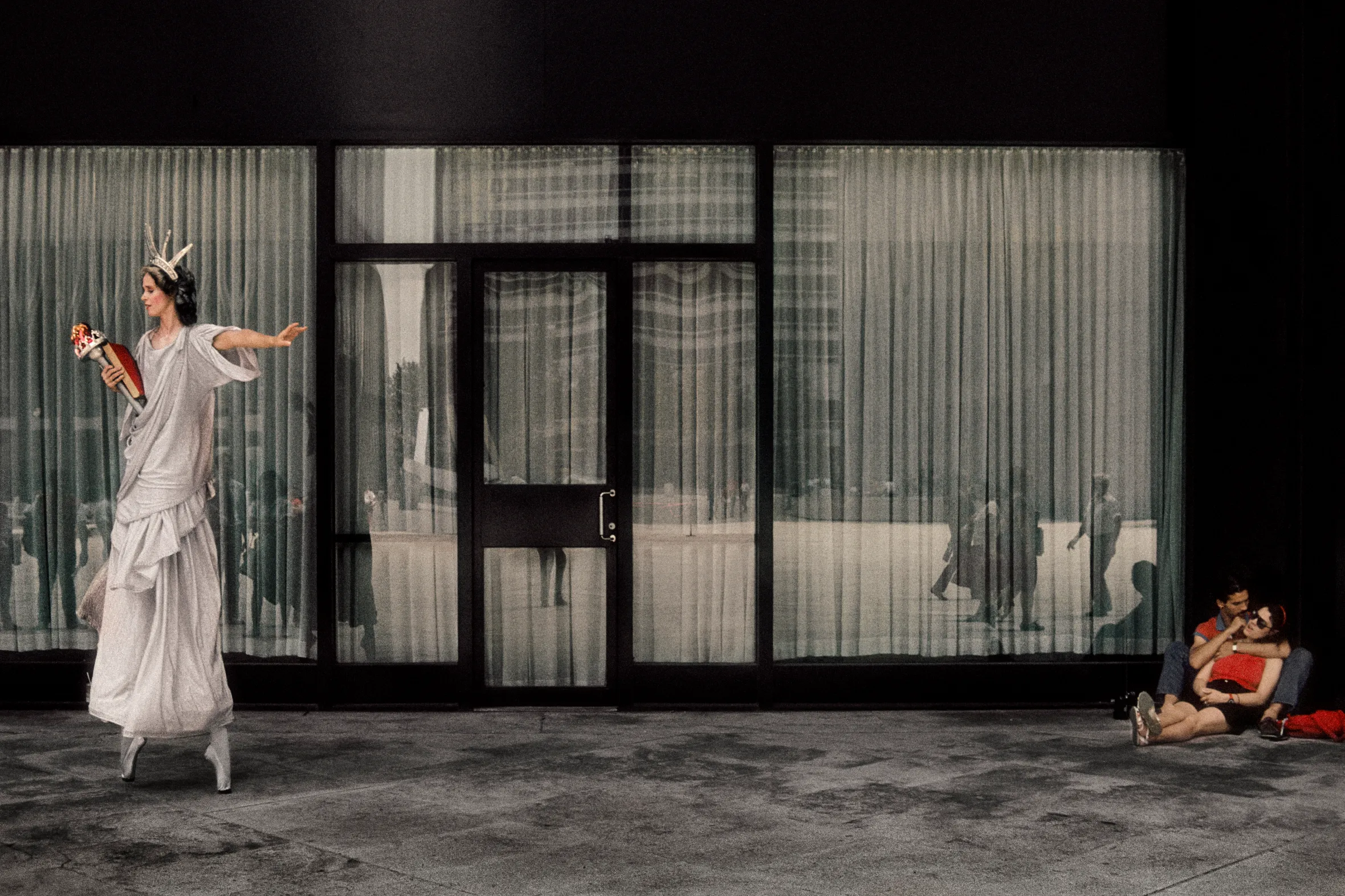 A woman dressed as the Statue of Liberty on pointe, holding a torch, is on a street in front of a building with large glass windows reflecting people walking by, while a couple sits on the ground to the right.