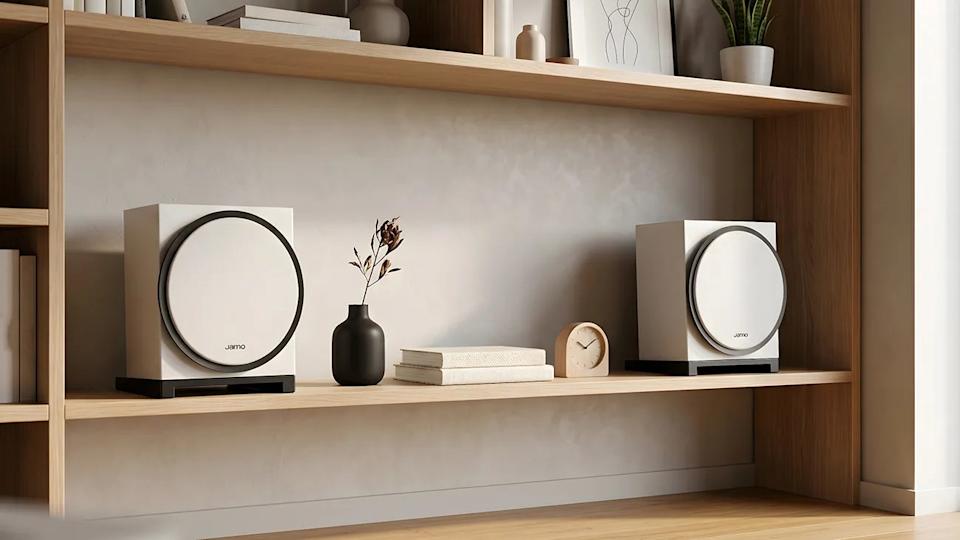 Two white Jamo speakers on a wooden shelf with a black vase, stacked books, and a small clock.