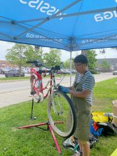 A person wearing an apron and gloves repairs a bicycle mounted on a stand under a blue canopy at an outdoor setup, with tools and bags on the ground nearby.