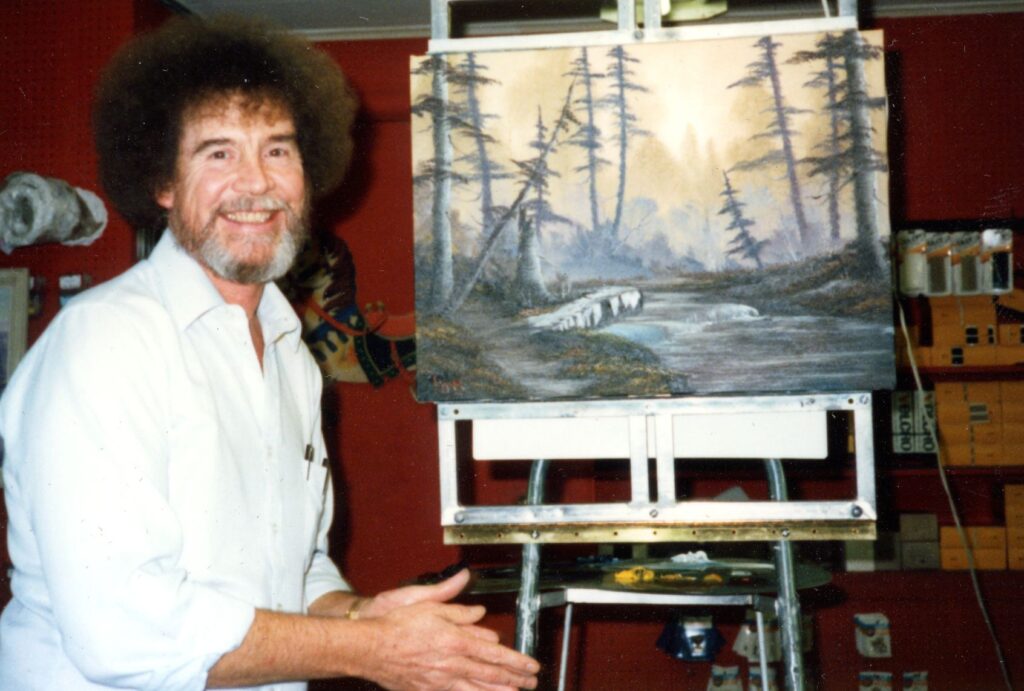 A smiling bearded man with an afro and wearing a white button-down shirt sits besides a landscape painting that includes skinny evergreen trees bordering a stream