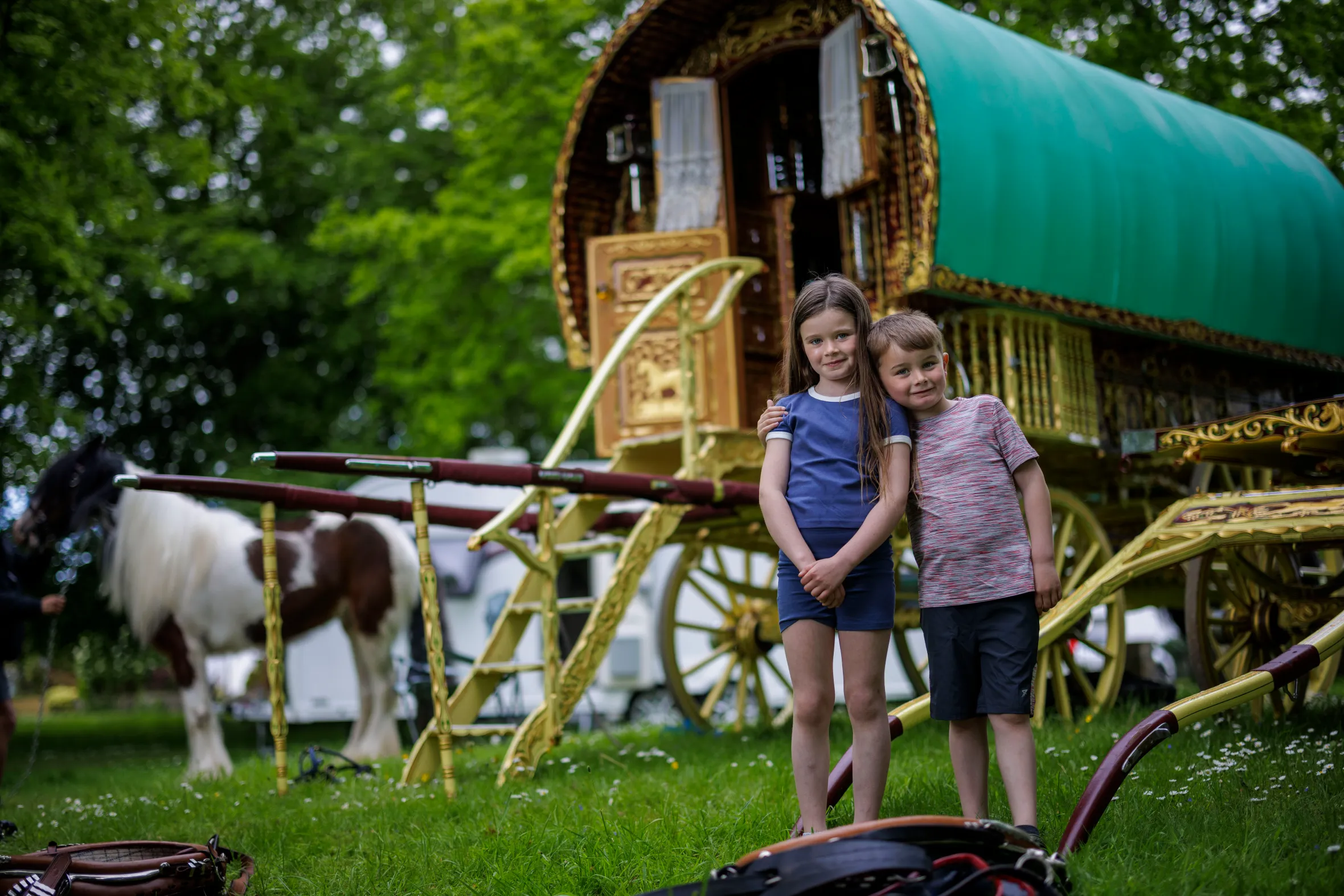 Two children, a girl and a boy, pose in front of a traditional gypsy wagon and a horse.