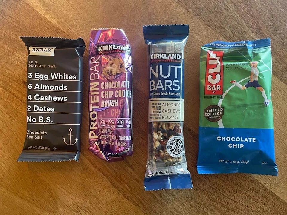 Four protein bars on a wooden surface: RXBAR, Kirkland Chocolate Chip Cookie Dough, Kirkland Nut Bar, and Clif Bar Chocolate Chip