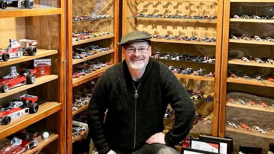 A man stands inside a room lined with wooden display cabinets filled with miniature racing cars. The shelves behind are densely arranged with detailed model vehicles in various colours and styles. The scene highlights a carefully curated collection showcased in a warm, museum‑like setting.