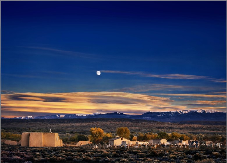 A fake-looking image of the moon in the sky above a western landscape, with a village in the foreground.