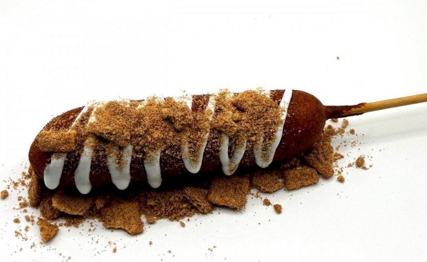 A deep-fried corn dog topped with sweet white frosting drizzle and crushed Cinnamon Toast Crunch cereal.