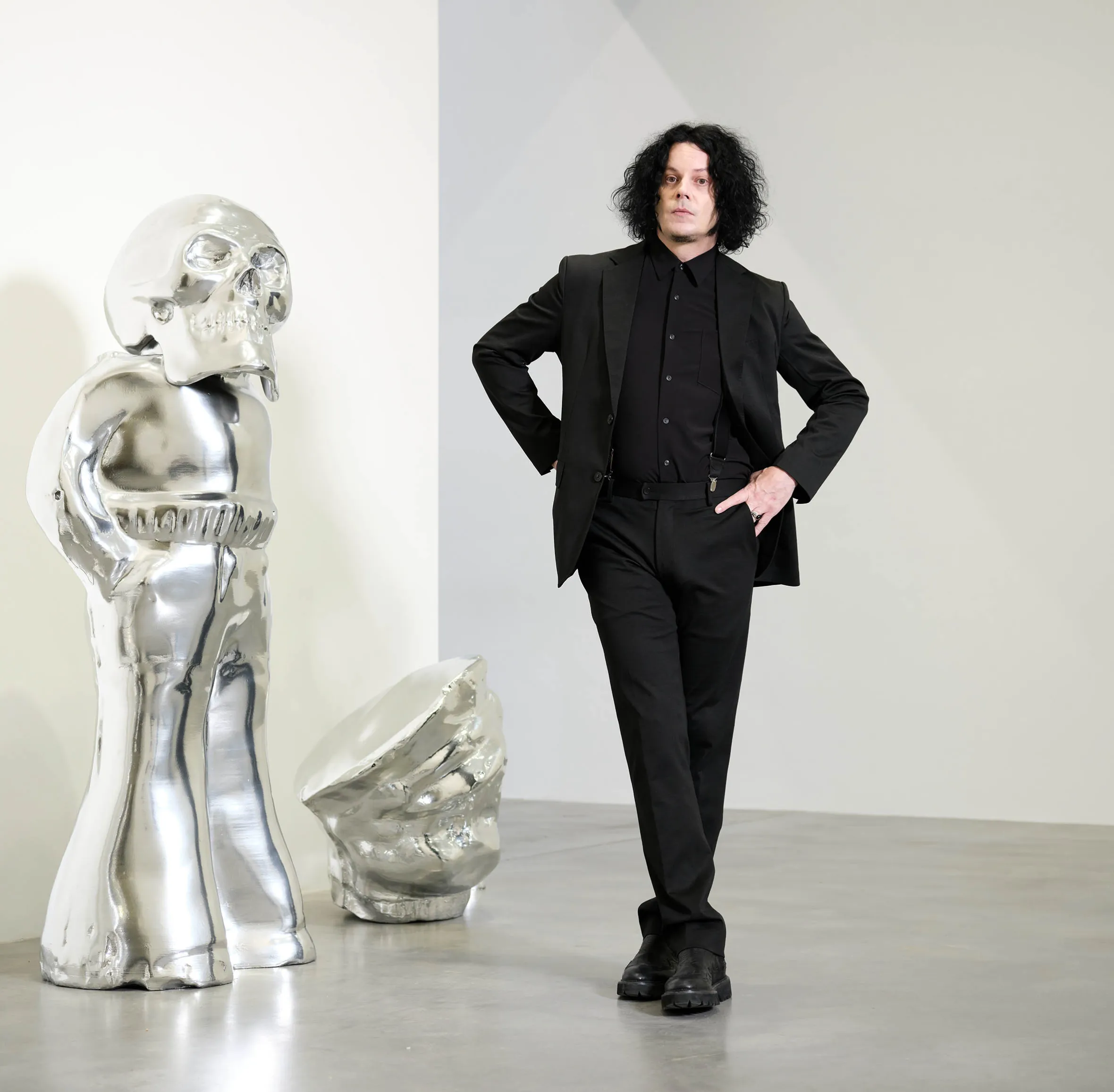 Jack White, an American artist, stands in a gallery next to two of his sculptures: a large, shiny, silver skull and another amorphous silver form.