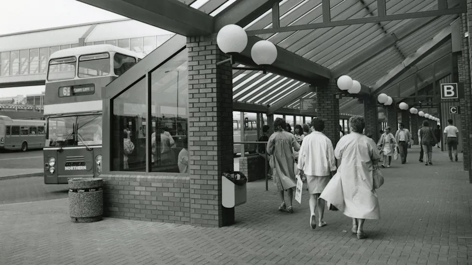 A black and white photograph of the bus station with a double decker bus on the right. On the right women dressed smartly are walking alway from the camera towards the stands. The woman on the right is wearing a trench coat and the woman next to her on the left is wearing a pencil skirt and jacket.