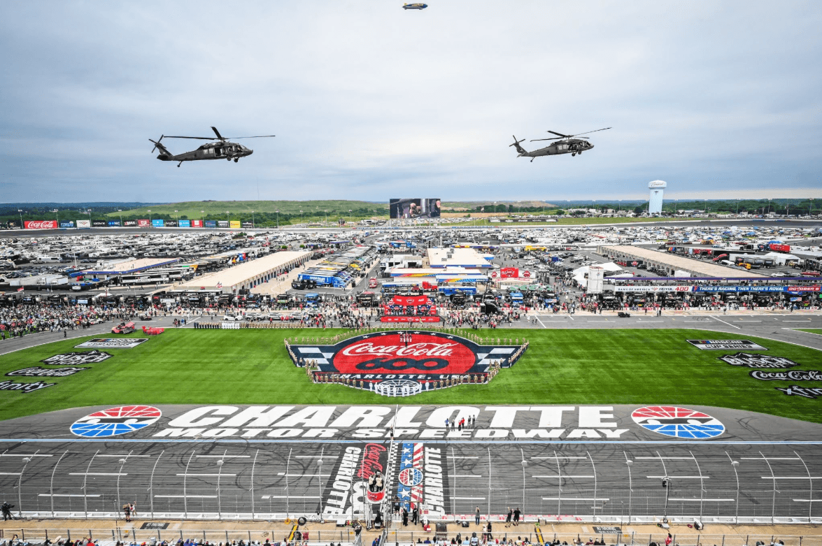 Aerial view of Charlotte Motor Speedway during a large Coca-Cola 600 event, with a green infield, crowds, and two helicopters flying low over the track.