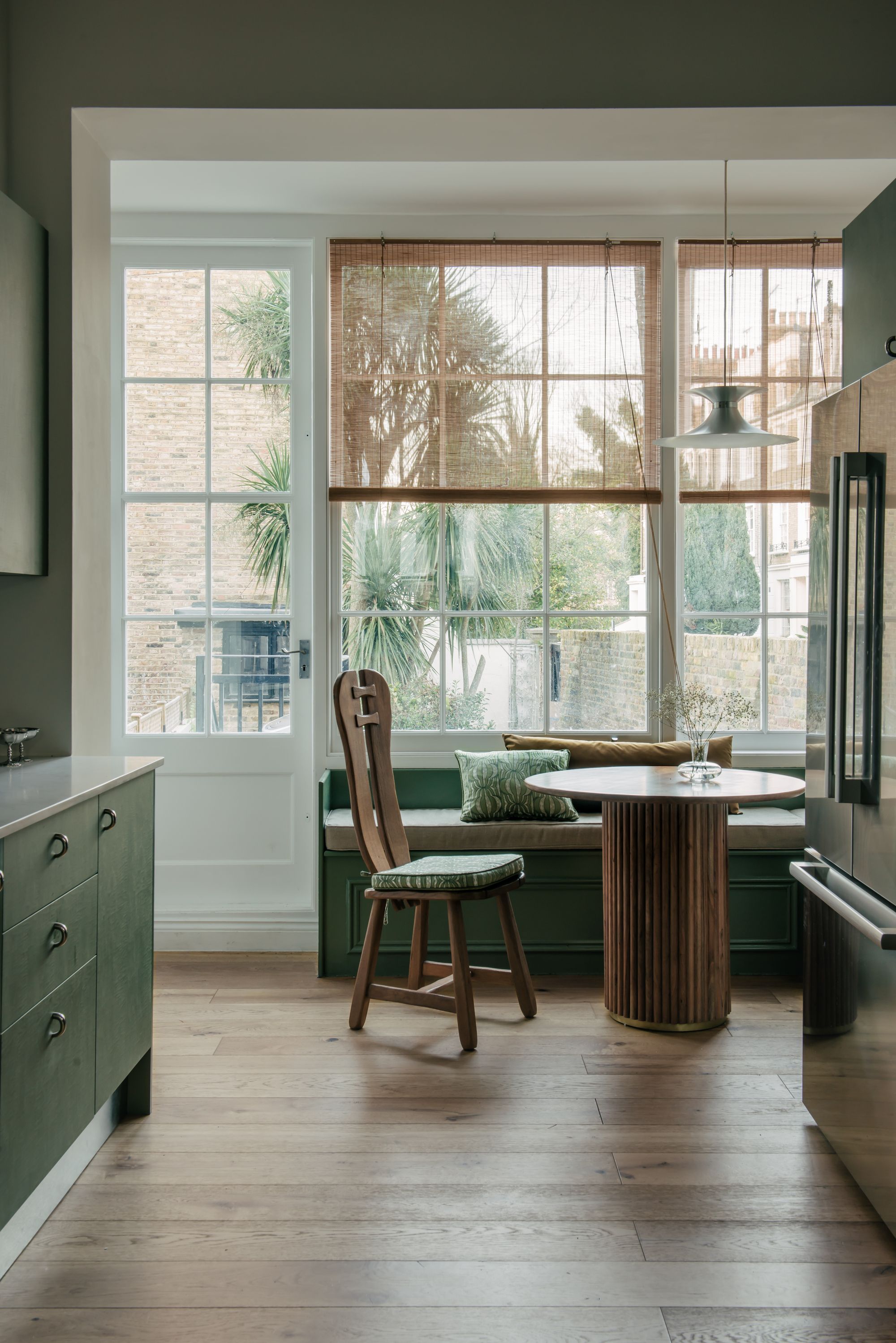 Image of a bright breakfast nook with lots of windows off of a kitchen. The space is decorated in a green color scheme and there are bamboo blinds and a wooden table.