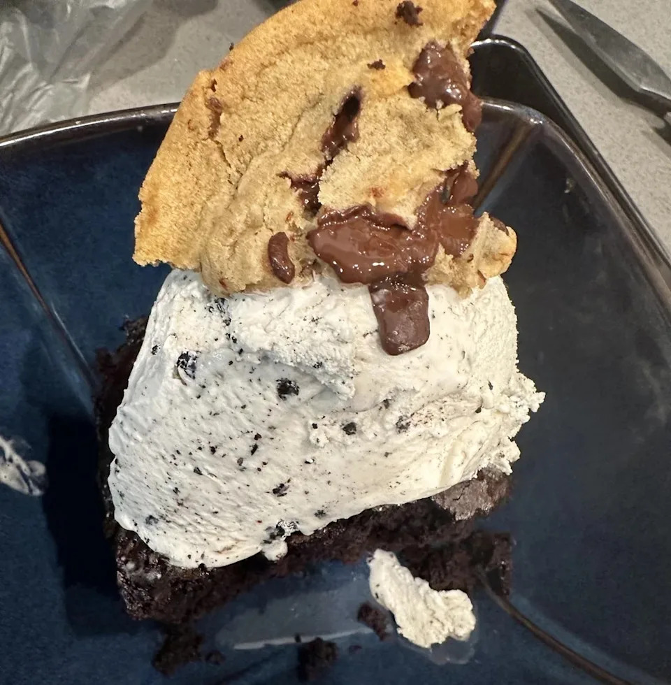 A decadent dessert with a scoop of cookies and cream ice cream on chocolate cake, topped with a chocolate chip cookie