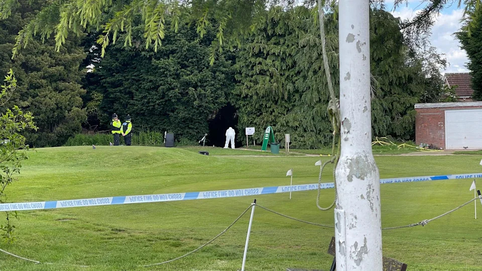 Police tape at the golf course behind a tree. Figures can be seen in the distance around a path.