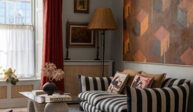 Living room with pale blue walls, striped sofa, red curtains, a glass and stone coffee table, a large painting above the sofa in orange and red of houses, and a rattan floor lamp in the corner