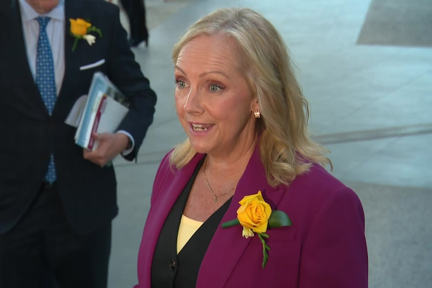A blonde woman wearing a magenta blazer and a yellow flower.
