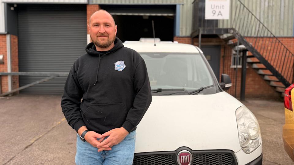 A man with short hair and beard, wearing a black hoodie and blue jeans. He is perched on the bonnet of a white fiat van.