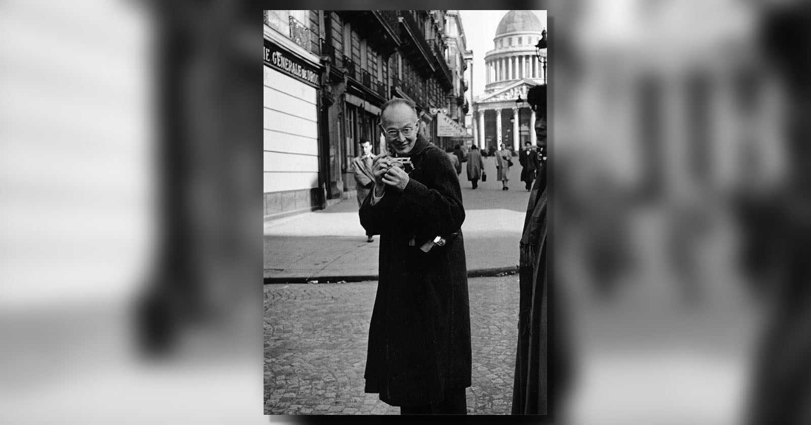 A man in a dark coat smiles while holding a cat on a city street. In the background, pedestrians walk near classical buildings, including a domed structure. The scene appears to be in an urban European setting.