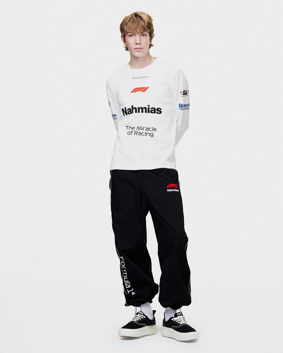 A person with light brown hair wearing a white long-sleeve shirt and black pants with "Nahmias" and "Formula 1" branding, standing against a plain background.