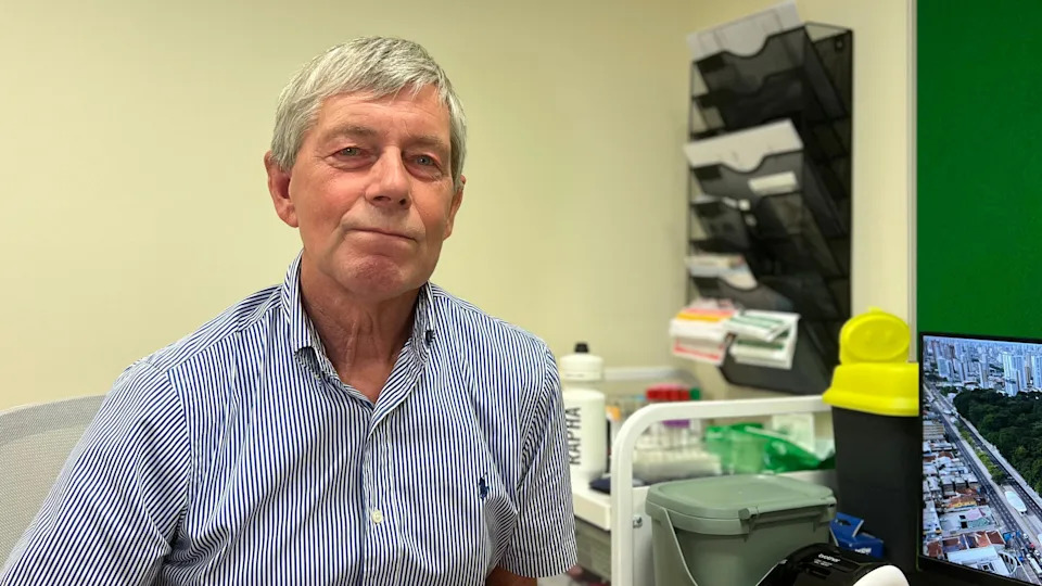 A person with short grey hair wearing a blue-and-white striped shirt sits in a clinical office setting, with shelves, trays, and medical supplies visible in the background alongside a computer monitor.