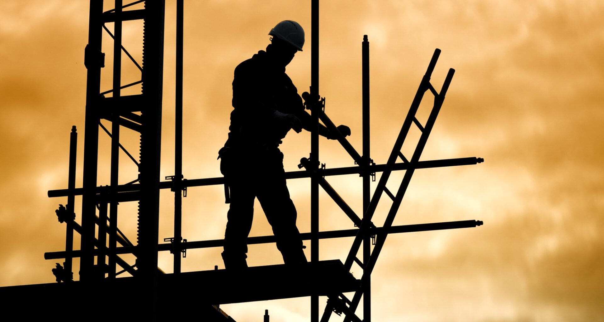 Silhouette of a construction worker on scaffolding, against a dramatic golden sky. The scene conveys industry and focus at dusk.