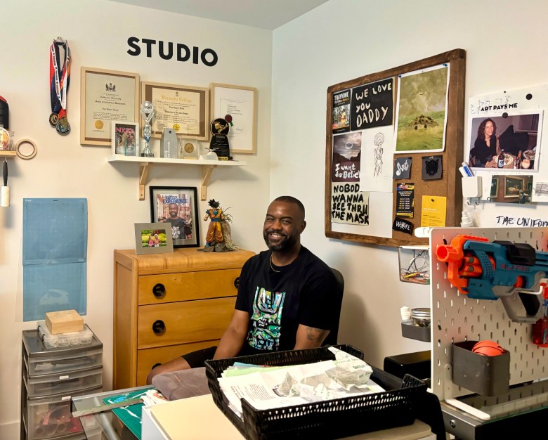 A smiling Black man wearing a black T-shirt with a vibrant print on the front sits behind a desk. Behind him on the wall in black lettering it says STUDIO, and a cork board to the right of the frame has a number of things tacked to it, including a note that says "WE LOVE YOU DADDY" and "I want to believe."
